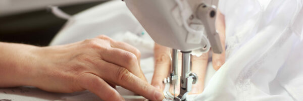 Close up view of sewing process. Female hands stitching white fabric on professional manufacturing machine at workplace. Seamstress hands holding textile for dress production. Light blurred background; Shutterstock ID 1416331862; Purchase Order: -